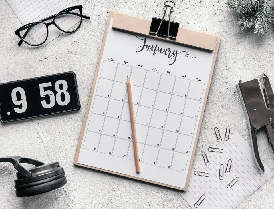 A paper calendar, digital clock and stationery on a desk.