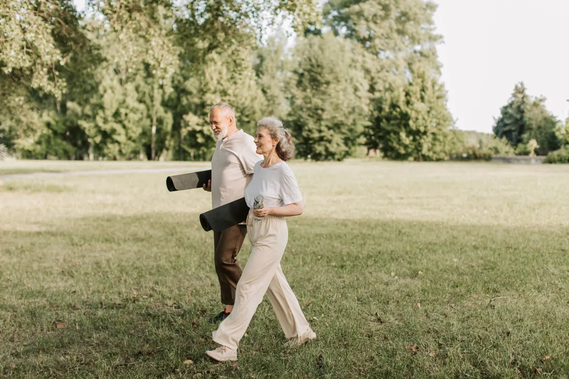 An older couple walking across grass while carrying rolled exercise mats.