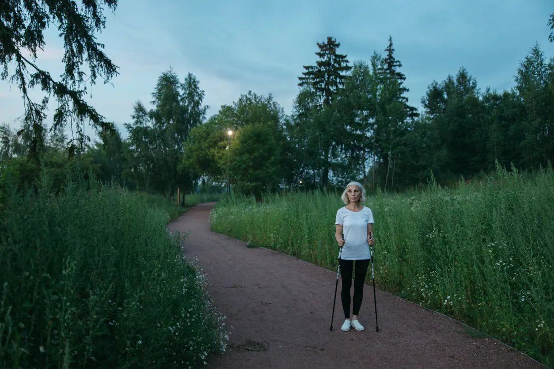 An older woman holding walking poles on a tree-lined park path.