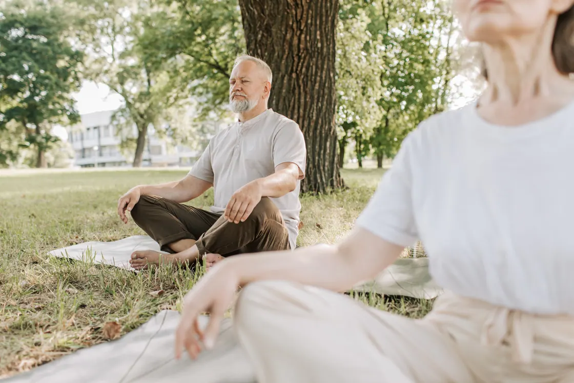 An older man sitting cross-legged on a mat beside another person in a park.