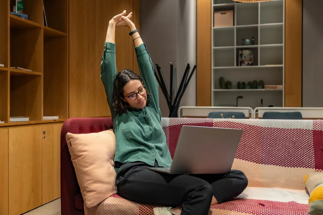 A woman stretching her arms overhead while sitting on a sofa with a laptop.