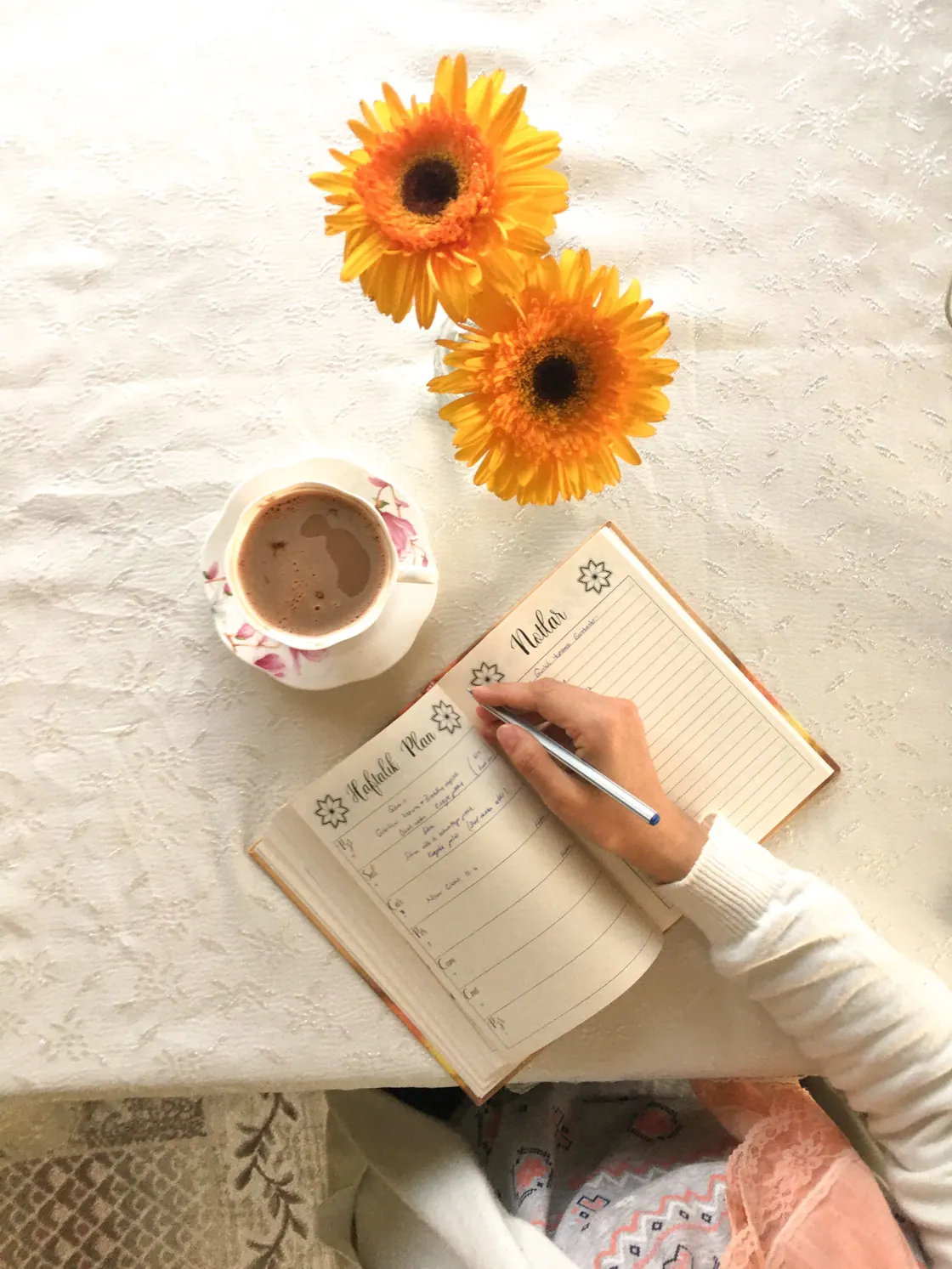 A hand writing in a planner beside a cup and two orange flowers.