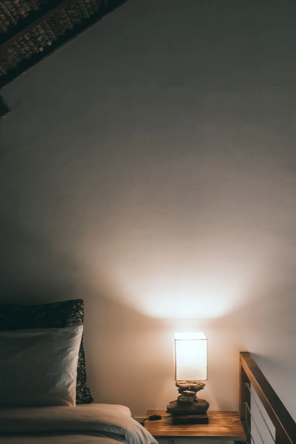 A lit bedside lamp beside pillows and a wooden bed frame.