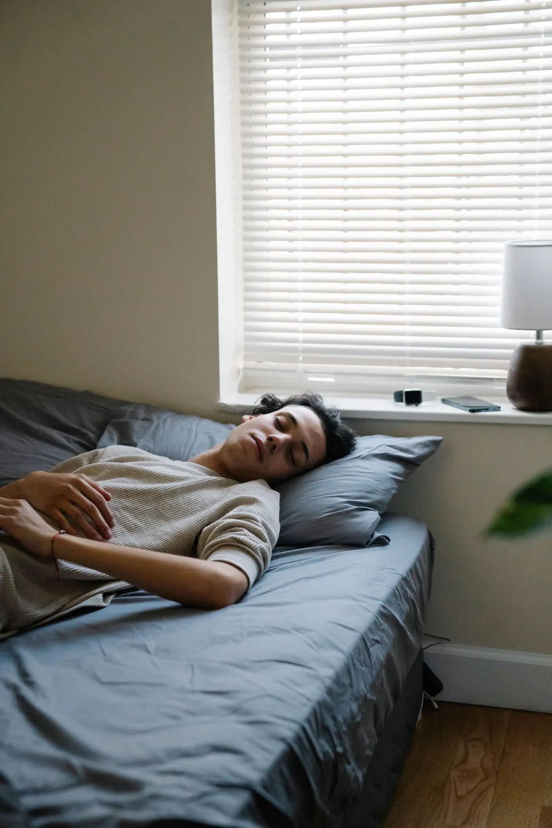 A man resting on a bed with gray bedding beside a window with blinds.
