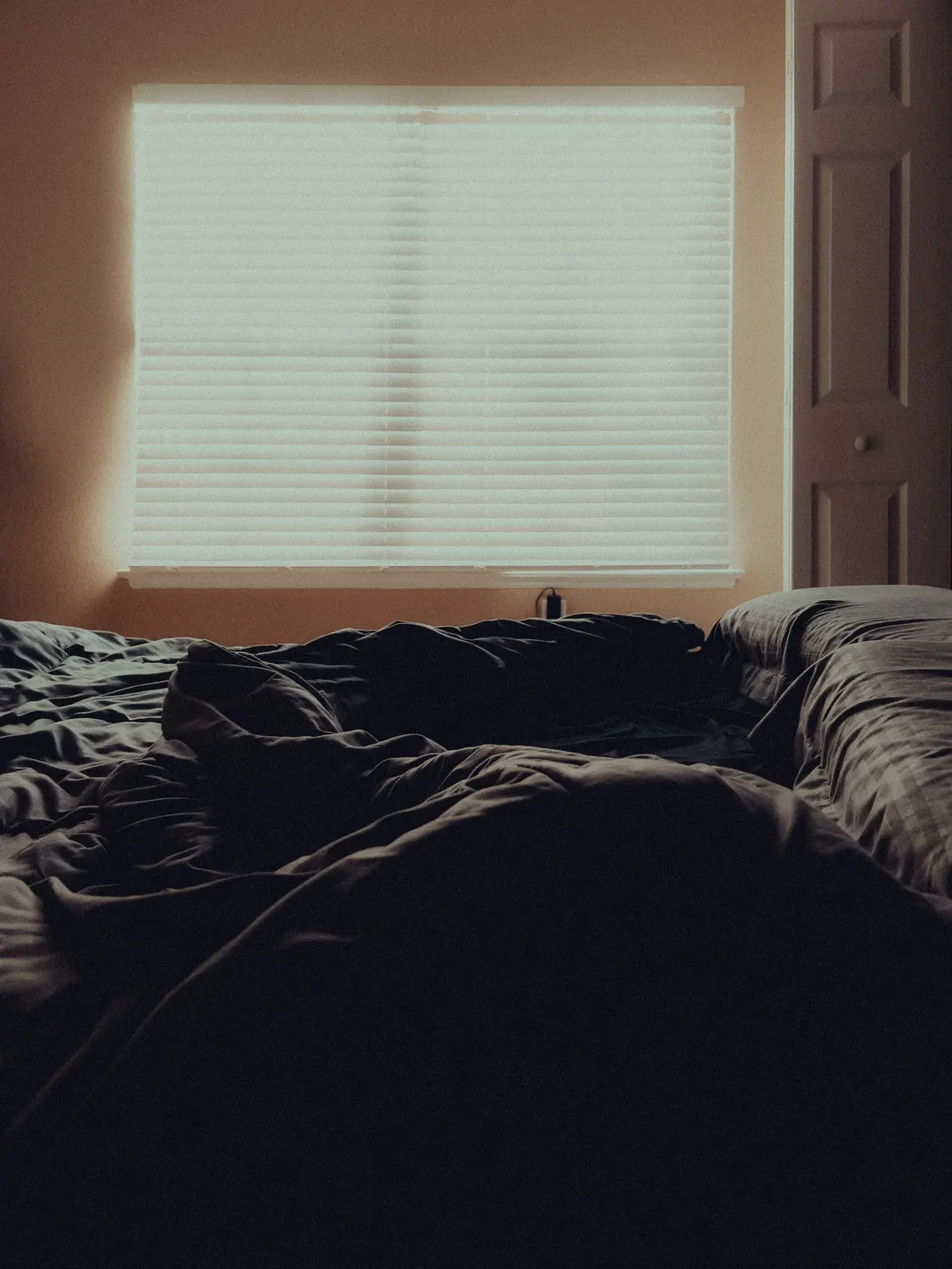 An unmade bed in a dim room with daylight filtering through window blinds.