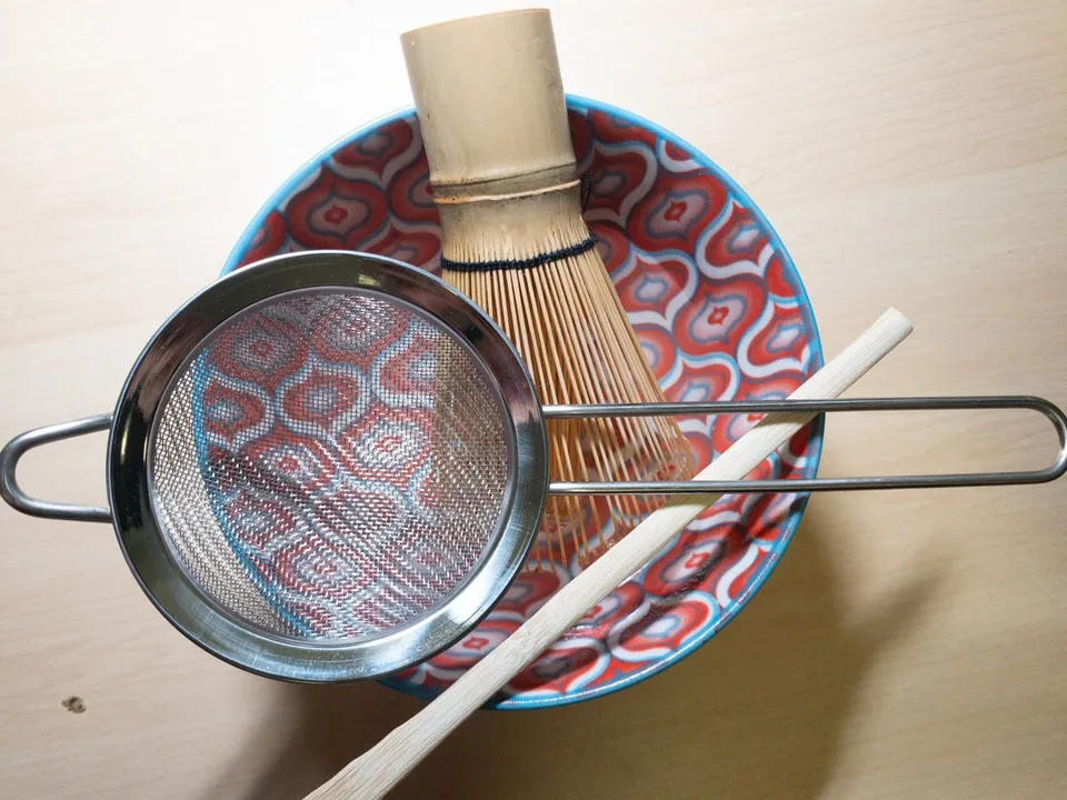 A bamboo whisk, bamboo scoop and metal strainer resting over a patterned bowl