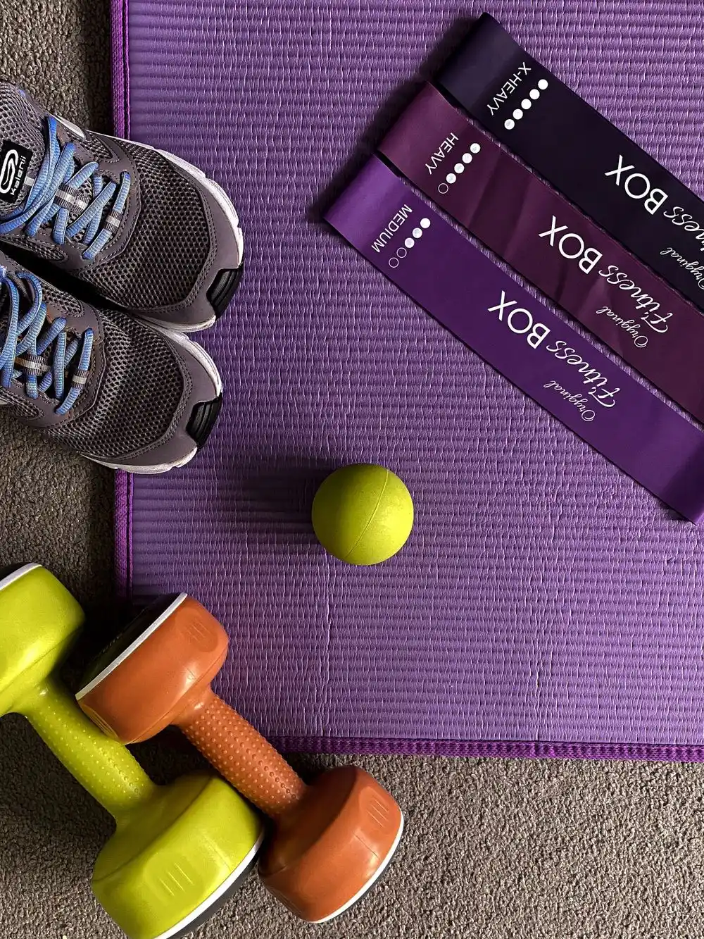Shoes, resistance bands, a ball and dumbbells arranged on a purple exercise mat.