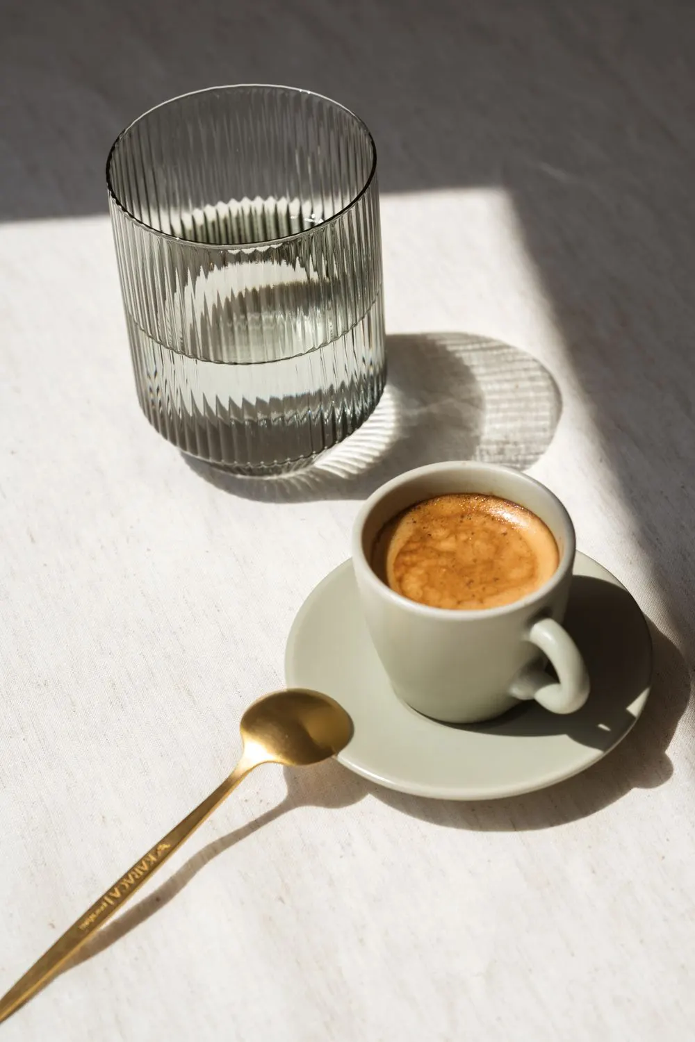 A cup of espresso and a glass of water on a sunlit table.
