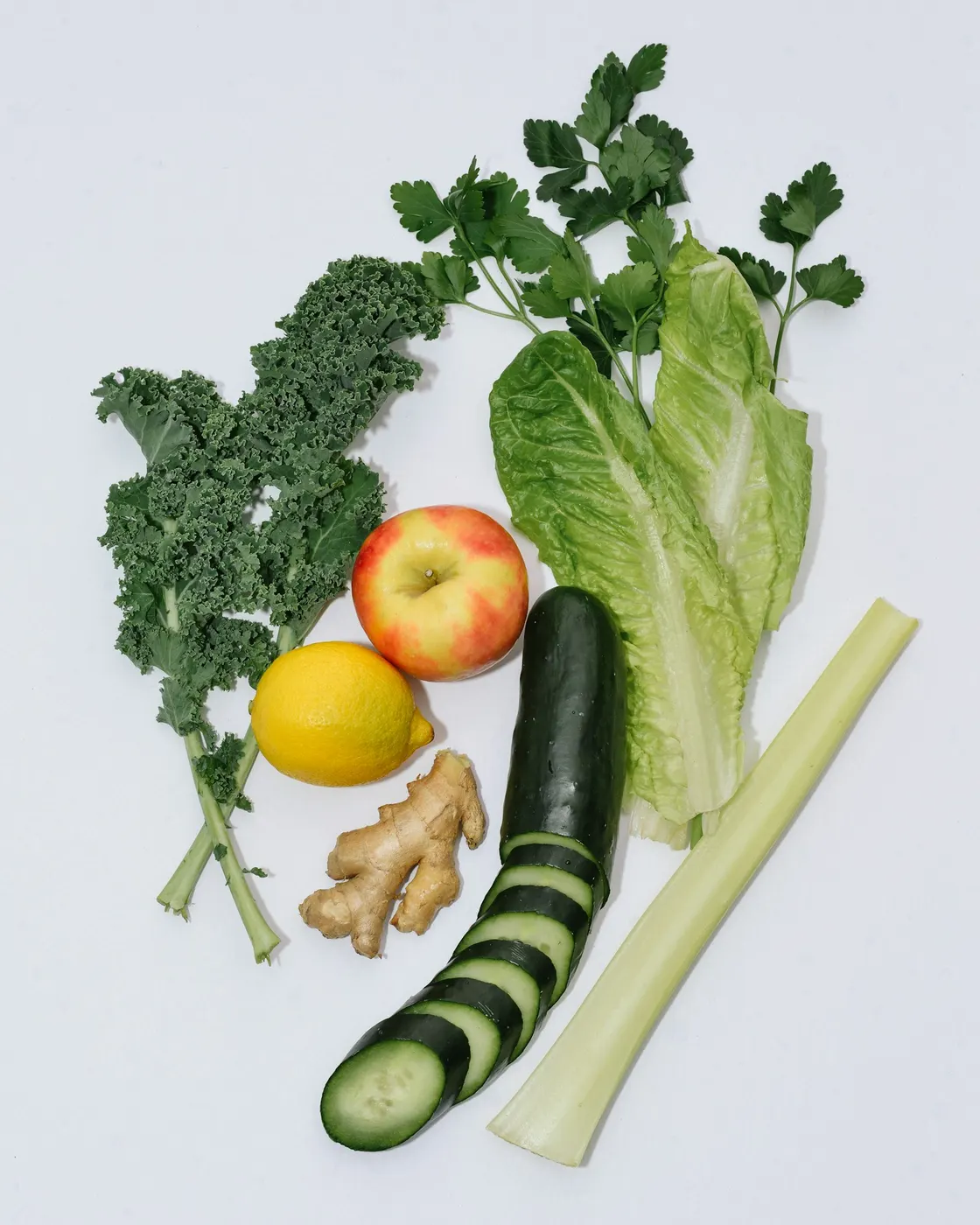 Leafy vegetables, cucumber, celery, ginger, an apple and a lemon arranged on a light surface.
