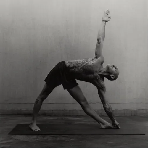 A person standing with feet wide apart, one hand resting near the front ankle and the opposite arm extended upward.