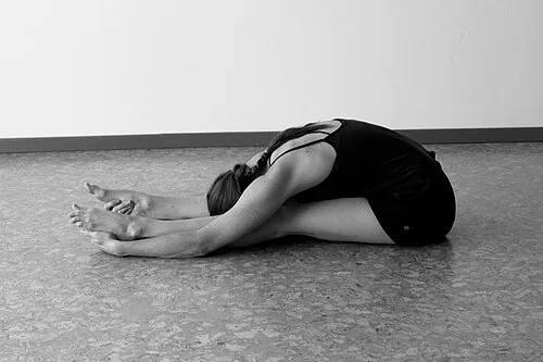A person seated on the floor with legs extended, folded forward with the head lowered, in a black-and-white photograph.