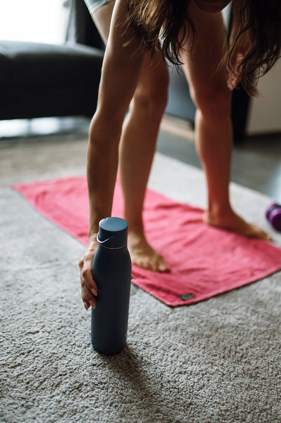 A person leaning forward to pick up a dark water bottle beside a pink exercise mat on the floor.