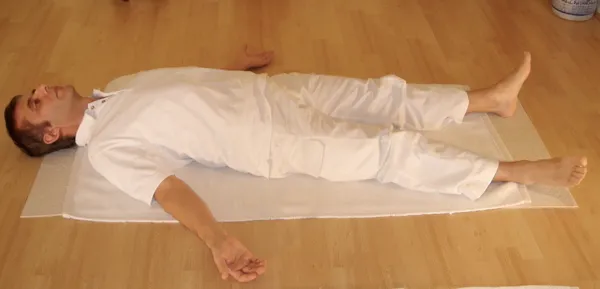 A person in white clothing lying on their back on a mat, with arms resting away from the torso and legs extended.